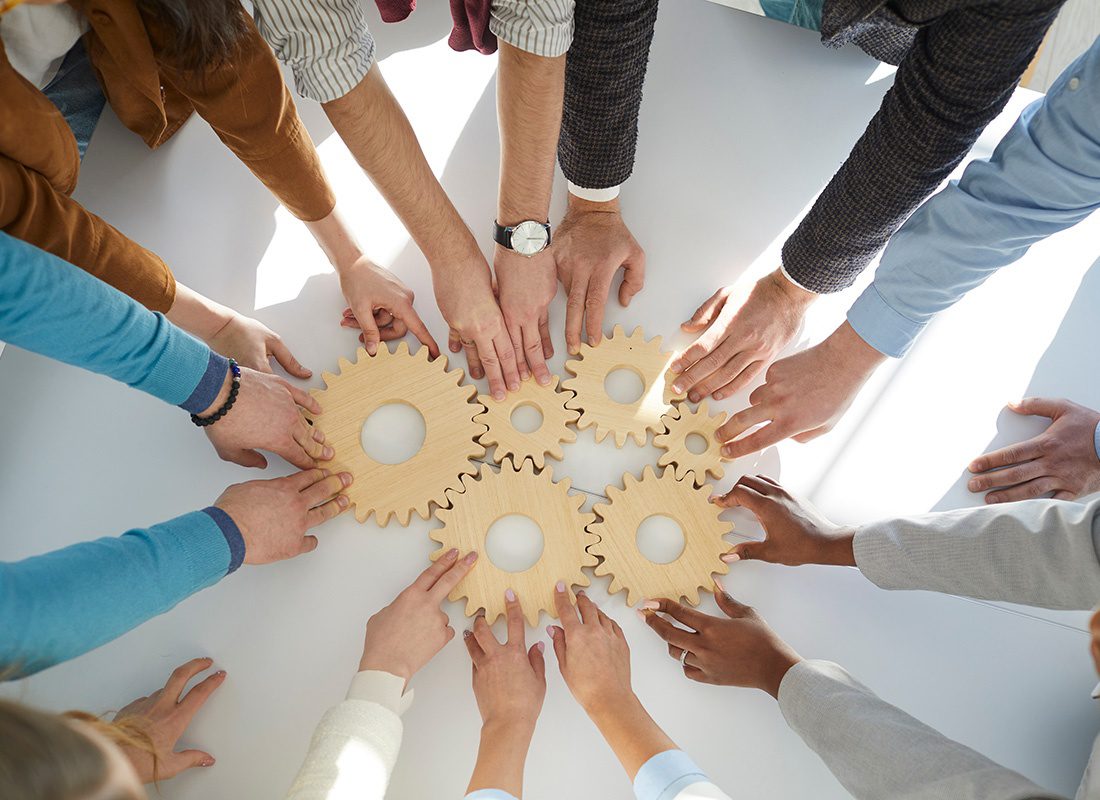 Employee Benefits - View of the Hands of a Group of Employees Standing in a Circle Holding Multiple Wooden Cog Pieces on a Table