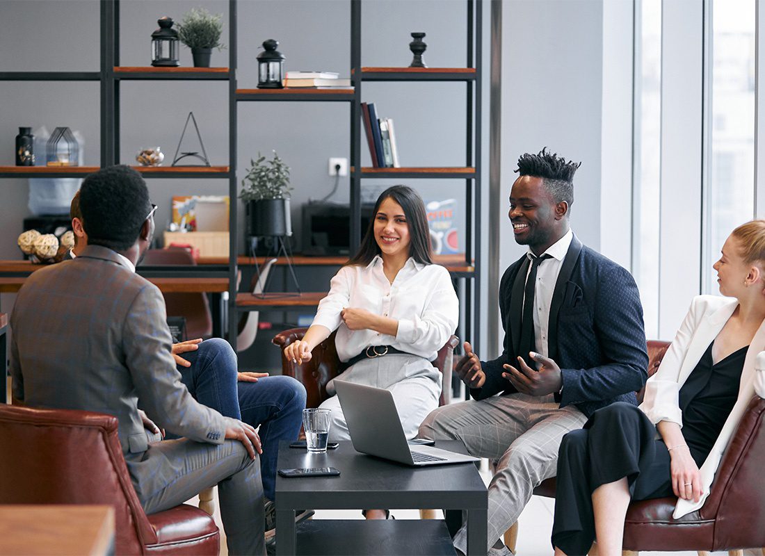 Group Benefits - View of a Cheerful Group of Employees Sitting Around a Coffee Table with a Laptop During a Meeting in a Modern Office Room Having a Discussion