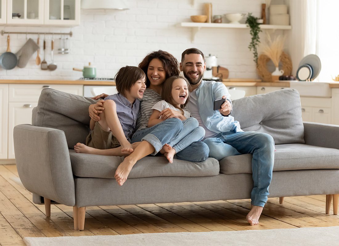 Personal Insurance - Portrait of Cheerful Parents Having Fun Spending Time with Their Two Kids While Sitting on the Sofa in the Living Room and Watching Videos on a Phone